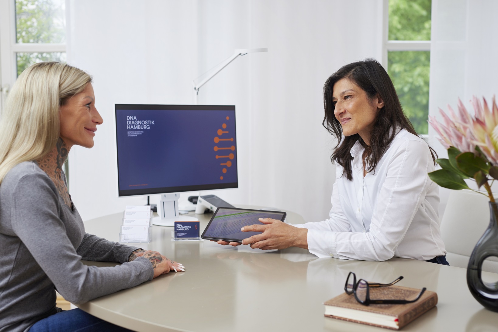 Humangenetikerin hält ein iPad und bespricht die Laborergebnisse mit einer Patientin im Arztzimmer